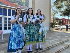 A 14ª edição da Festa do Vinho de Santana, tradicional evento da colônia tirolesa de Piracicaba, acontece neste feriado prolongado, de 20 a 22 de junho, na praça Bortolo Vitti, com programação que inclui apresentações artísticas, abrangendo música e dança, além de pratos típicos. O evento é realizado pela Associação de Moradores de Santana e Circolo Trentino di Piracicaba, com apoio da Prefeitura, por meio das secretarias municipais de Turismo e de Cultura. A entrada é gratuita. As informações sobre a festividade foram divulgadas nesta terça-feira, 17/06, em coletiva de imprensa com a presença dos organizadores e autoridades. Na sexta, 20/06, a abertura oficial será às 20h, com apresentação da rainha Camilly Moraes Forti, a princesa Gabriela Vitória Rodrigues e a embaixadora Juliana Biason Vitti. Na sequência, às 21h, as atrações serão grupos de dança, seguidos, às 22h, de show com a banda Elas Cantam Italianas. No sábado, 21/06, a festa tem início às 19h30, também com apresentação da rainha, da princesa e da embaixadora. Na mesma noite, às 20h, grupos de dança animam o evento. E às 22h, haverá show da banda Nostalgia. Para o último dia, domingo, 22/06, o público poderá participar, às 11h, do almoço com prato colonial, composto por arroz, crauti (repolho curtido refogado, pimenta, sal, costelinha de porco defumada, bacon e calabresa), strongolapreti (massa feita com espinafre ao molho vermelho) e porpeta. Quanto às atrações artísticas, haverá banda itinerante, apresentação da realeza às 14h e show com a banda Nostalgia às 15h. Na sexta e no sábado, a festa termina à 1h (do dia seguinte). No domingo, às 19h. Neste ano, os vinhos disponíveis na festa são da vinícola Alma da Videira, produzidos no próprio bairro. Entre os destaques estão o Vinho do Nono (uva bordô), nas versões seco e suave, além do vinho branco (uva Lorena). As novidades são o vinho tinto seco Merlot e o suco de uva integral. O cardápio também tem o canederli, feito com pão, linguiça, ovos e queijo, resultando em uma massa em formato de pequenos bolinhos cozidos em um caldo de frango, e a cucagna, que na receita tem linguiça, ovos, bacalhau e queijo, fritos separadamente e misturados na polenta. E a novidade do menu deste ano é pizza. A imagem mostra uma mesa coberta com uma toalha xadrez vermelha e verde, com vários itens dispostos sobre ela. Em primeiro plano, há uma caixa de madeira contendo vários pequenos doces embalados individualmente em sacos plásticos transparentes. Ao lado da caixa, há dois recipientes de plástico transparentes contendo uma sobremesa roxa escura, possivelmente um pudim ou mousse. Ao fundo, há uma variedade de garrafas de vinho e suco de uva. Há três garrafas de vinho tinto, uma garrafa de vinho branco e duas garrafas de suco de uva, todas com rótulos visíveis. O fundo da imagem é desfocado, mas mostra uma estrutura de vidro ou janela. A iluminação sugere que a foto foi tirada ao ar livre, em um dia ensolarado. A cena geral sugere uma mesa de piquenique ou uma apresentação de produtos alimentícios, possivelmente em um mercado de fazendeiros ou evento semelhante. Quitutes, vinho e suco foram apresentados durante coletiva de imprensa “Festas como esta são importantes para que se mantenham vivas as tradições e culturas de um povo. Além disso, atraem turistas e visitantes que, posteriormente, podem retornar ao município para aproveitar outros atrativos da cidade, ou seja, o movimento não apenas acontece no momento da festividade, ele pode refletir a longo prazo também”, comentou a titular da Secretartia de Turismo, Clarissa Quiararia. A imagem mostra um grupo de pessoas vestindo trajes tradicionais, possivelmente para uma celebração ou evento cultural. Há mulheres e meninas usando vestidos longos e coloridos, com detalhes que sugerem inspiração europeia, possivelmente alemã ou italiana. Duas das meninas usam faixas de concurso, indicando que podem ser rainhas ou princesas de alguma festividade. Homens, incluindo um padre, também estão presentes, vestindo roupas mais formais. O fundo é uma igreja, com um grande mosaico na parede que parece representar uma cena histórica ou geográfica. O ambiente parece ser externo, em um dia ensolarado. A foto sugere uma comemoração com forte identidade cultural e religiosa. Secretários municipais de Turismo e de Cultura, Clarissa Quiararia e Calros Beltrame, junto dos representantes do evento “É lindo ver o empenho da comunidade de Santana, que se une e se dedica no desenvolvimento desta festa, mantendo a tradição trentina tirolesa. O público com certeza percebe isso em cada detalhe do evento”, disse o secretário municipal de Cultura, Carlos Beltrame. Participaram da coletiva, também, Vera Lucia Negri, secretária do Circolo Trentino di Piracicaba, e o padre Joélio Brito, pároco da igreja Sant’Ana.